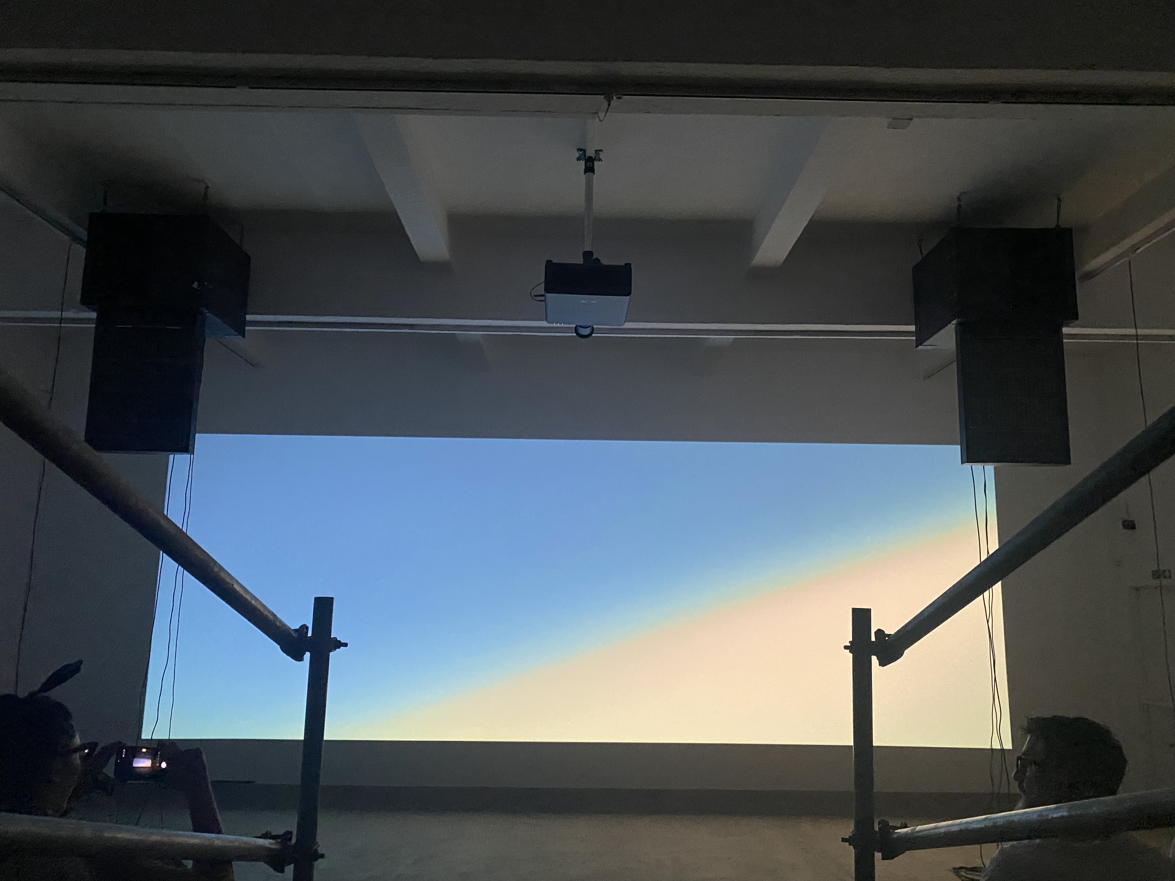 Dark gallery view of the projection wall and suspended audio hardware.